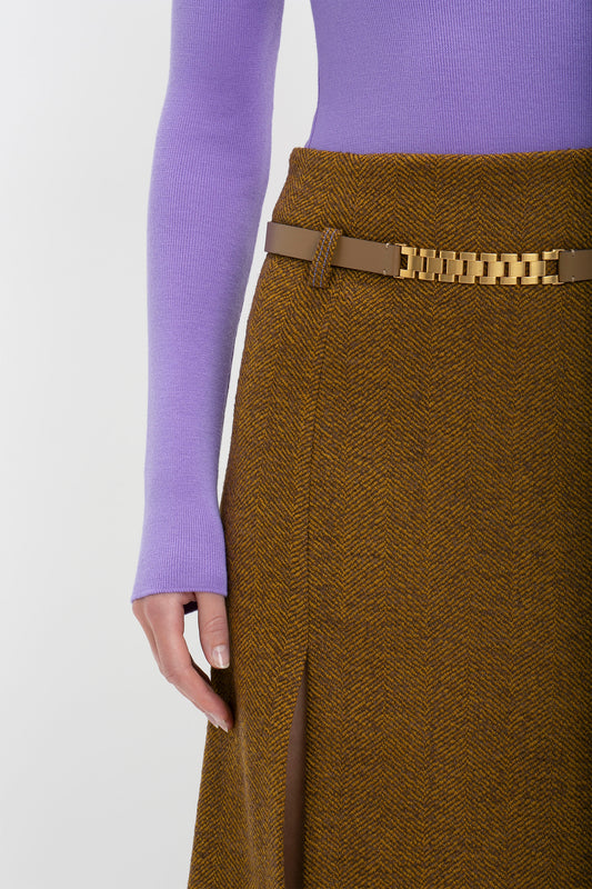 Close-up of a woman wearing a purple knitted polo neck jumper and a brown herringbone skirt with a Victoria Beckham UK Watch Strap Detail Belt in Khaki-Brown.
