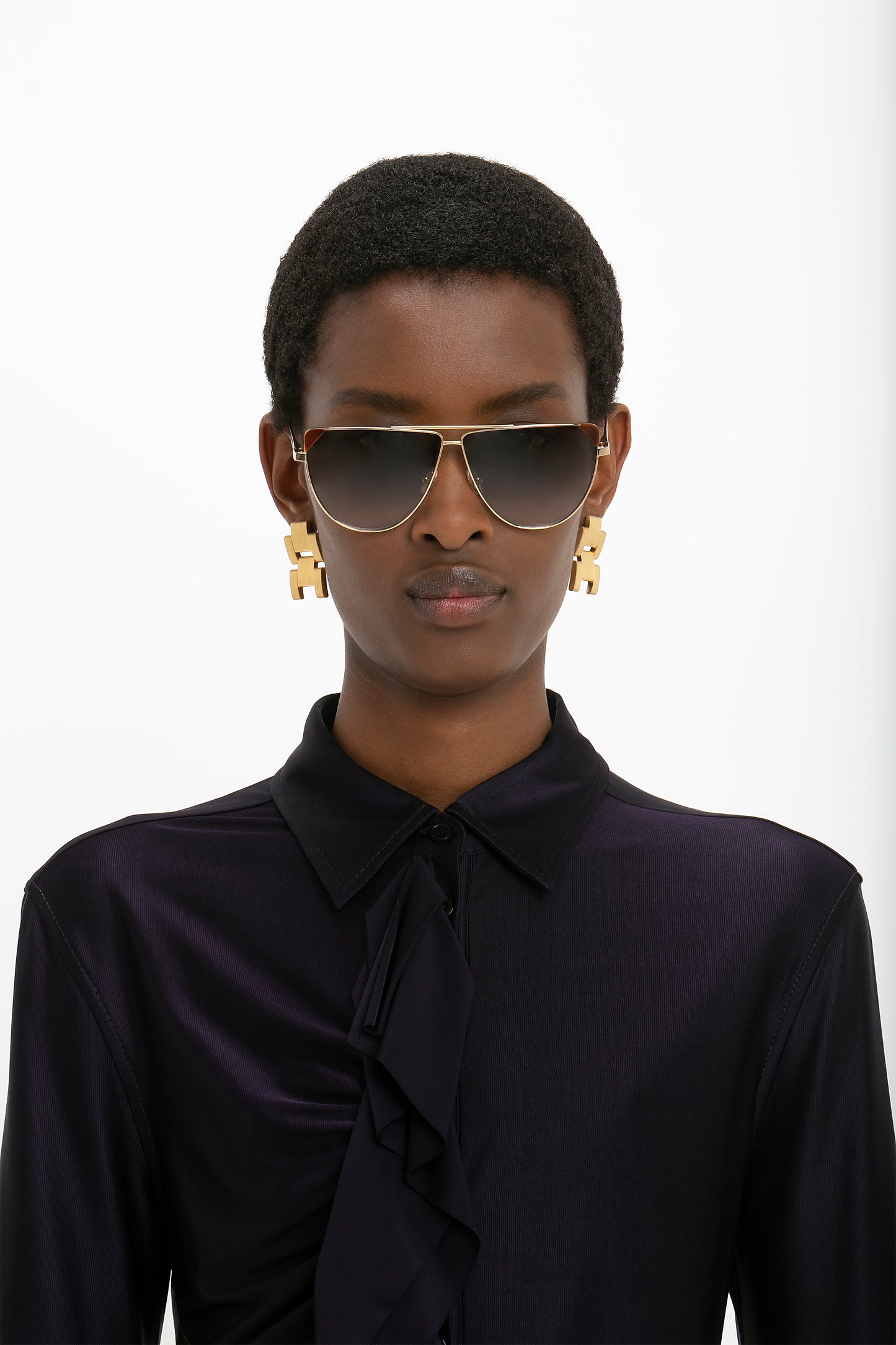 Person with short hair wearing Victoria Beckham V-Line Aviator In Gold Blonde Havana sunglasses, large gold earrings, and a dark, ruffled blouse against a plain background.