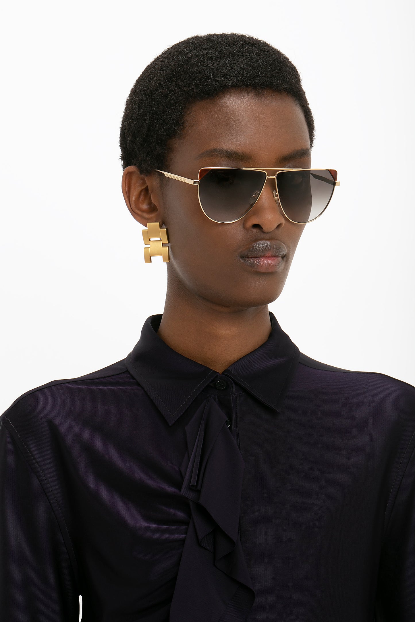 Person wearing a dark blouse, oversized gold earrings, and Victoria Beckham V-Line Aviator In Gold Blonde Havana sunglasses against a white background.