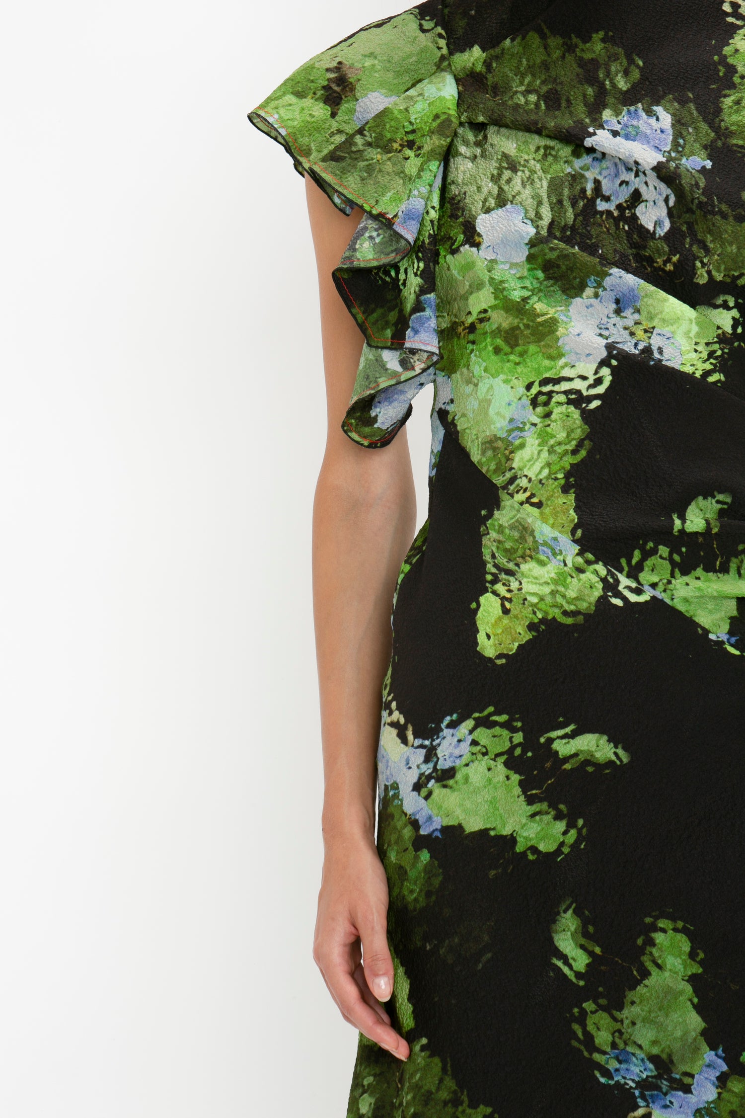 Close-up of a person wearing a black Twist Shoulder Dress In Black Frost by Victoria Beckham with green and blue abstract patterns, a single ruffled sleeve, and an asymmetric hemline. The image captures the waist-defining fit from the chest down to just above the knee.