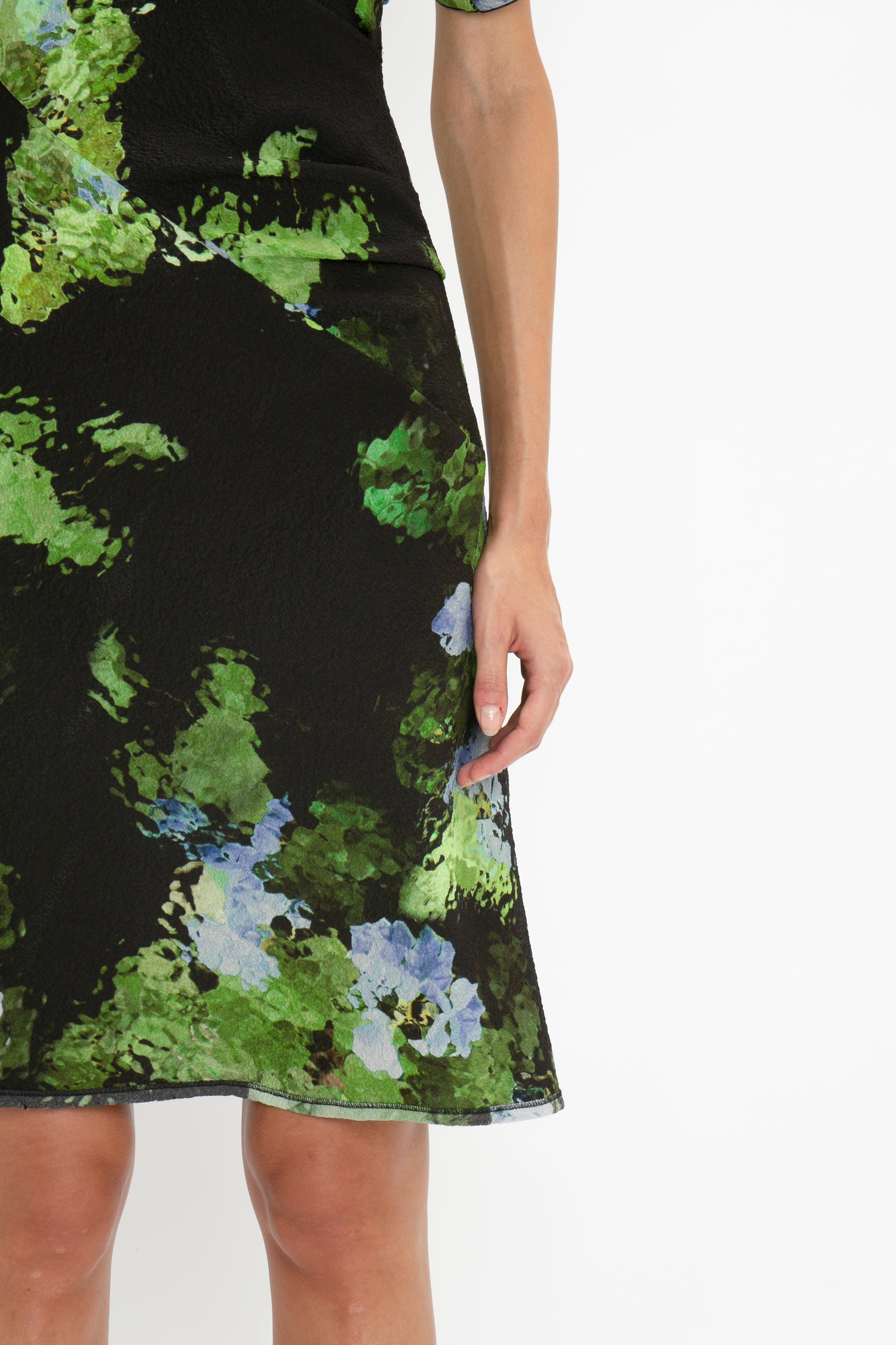 A person wearing a Twist Shoulder Dress In Black Frost by Victoria Beckham with green and blue floral patterns. Only the lower half of the waist-defining fit dress and part of their right arm are visible against a white background.