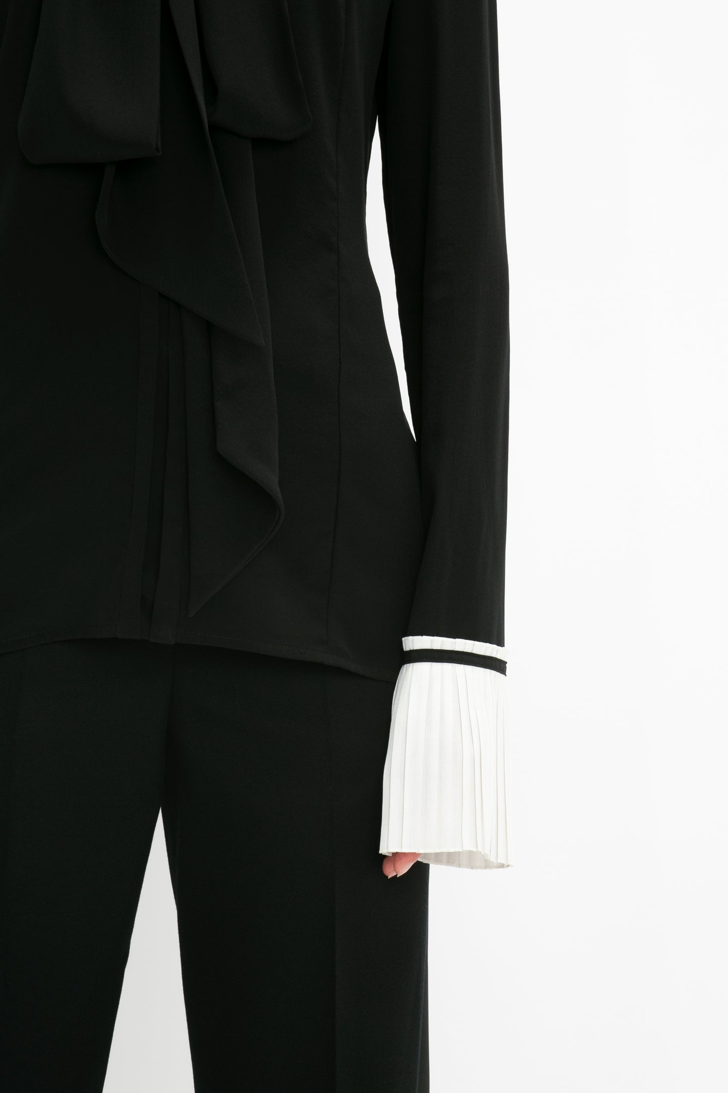 Close-up of a person wearing the Victoria Beckham Pleat Cuff Blouse In Black with an asymmetric ruffle detail and black pants. The white knife pleat cuff is visible on the left sleeve.