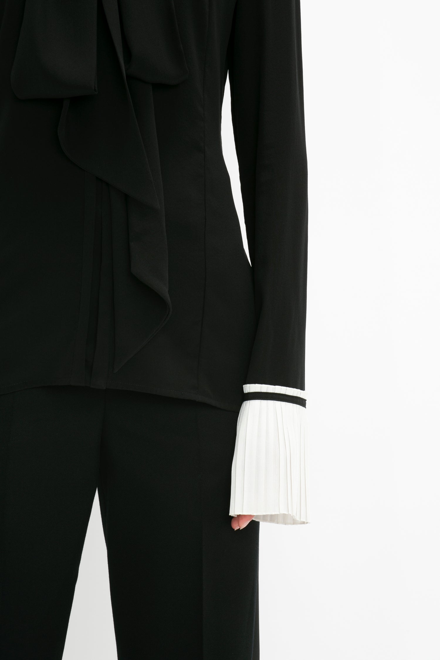Close-up of a person wearing the Victoria Beckham Pleat Cuff Blouse In Black with an asymmetric ruffle detail and black pants. The white knife pleat cuff is visible on the left sleeve.