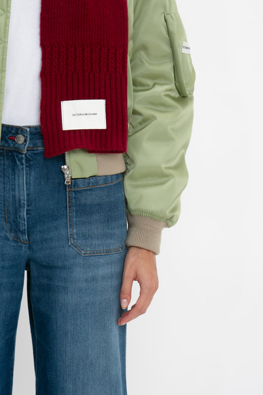 Close-up of a person wearing a green bomber jacket, Victoria Beckham Exclusive Logo Patch Scarf in Burgundy, and blue jeans, focusing on the midsection and left hand by the side.