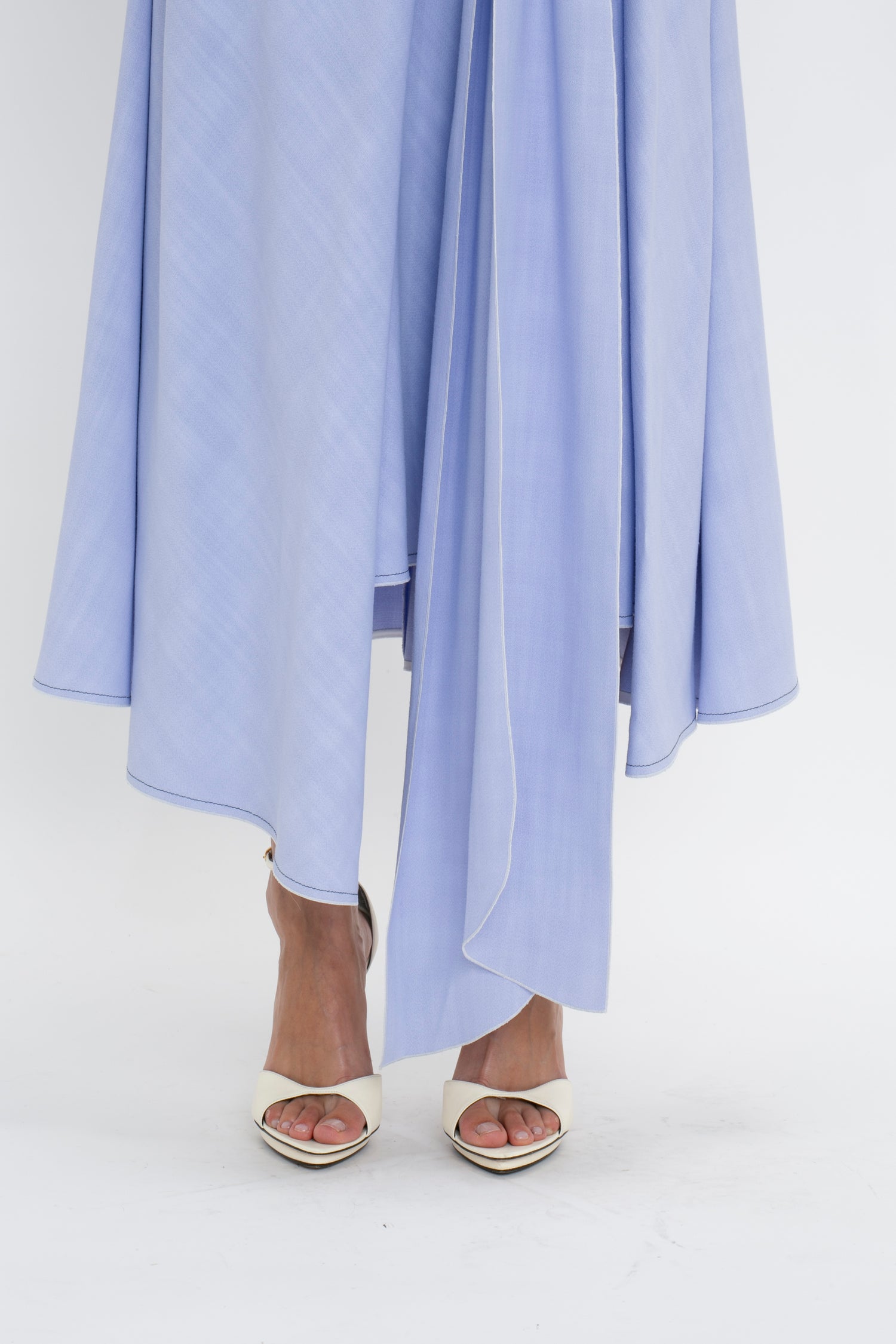 A person stands wearing cream-colored open-toe sandals and a light blue, Asymmetric Tie Detail Skirt In Frost crafted from fluid viscose crepe fabric by Victoria Beckham, with the image focused on their legs and feet against a white background, exuding modern femininity.
