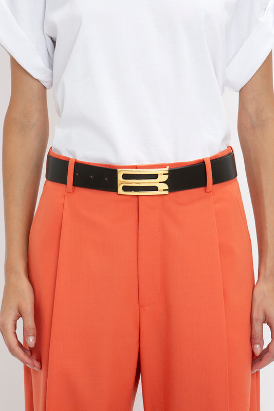 Close-up of a person wearing a white t-shirt and orange trousers with a Victoria Beckham Exclusive Jumbo Frame Belt In Black Leather featuring gold hardware.