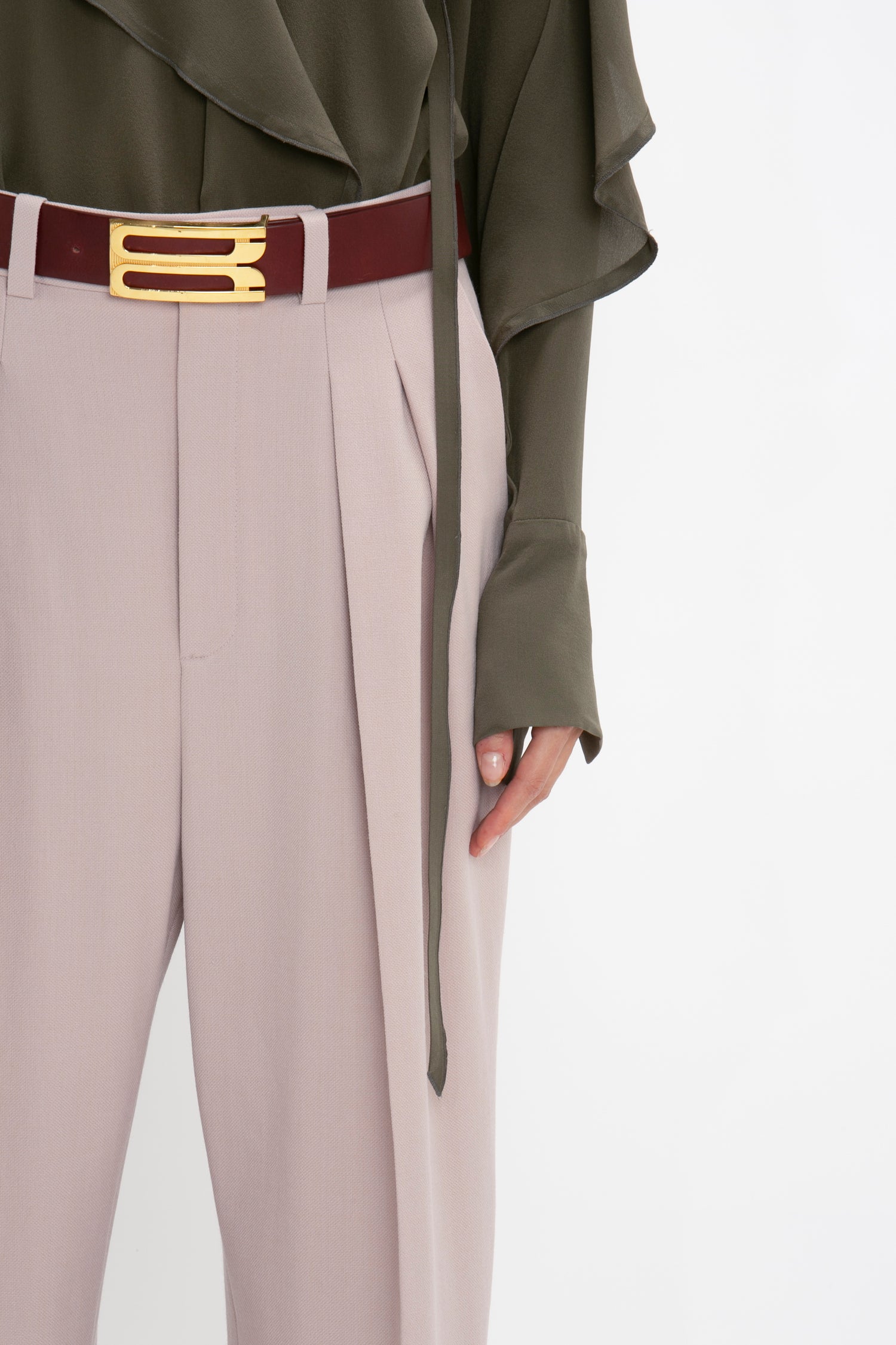 A person wearing beige double pleat trousers, a Victoria Beckham Tie Detail Ruffle Blouse in Oregano, and a red belt with a gold buckle stands against a white background.
