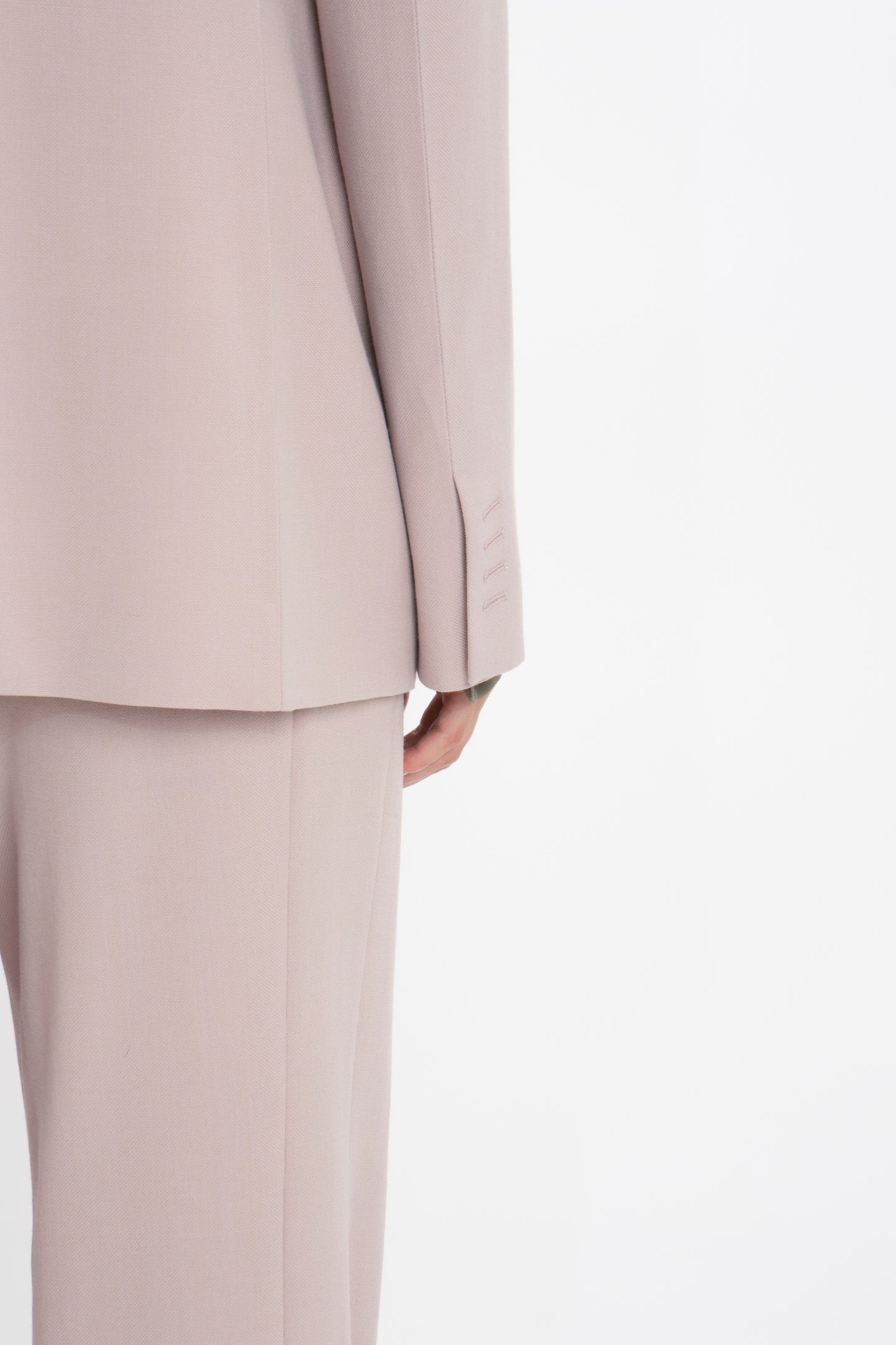 Close-up of a person wearing a light beige, menswear-inspired suit. The photo shows the back view of the Victoria Beckham Double Panel Front Jacket In Rose Quartz and a portion of the pants. Only the lower half of the body is visible, highlighting the exquisite fit and detailing.