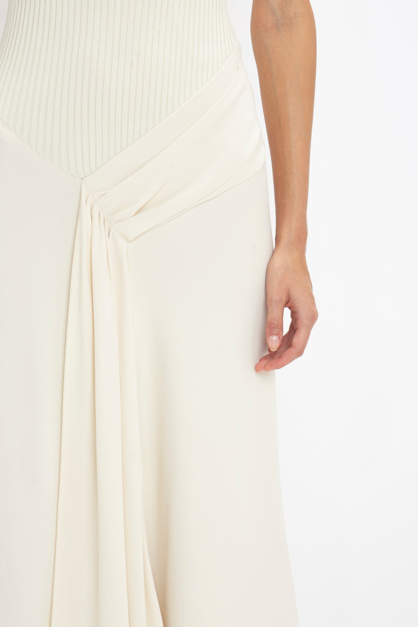 A close-up of a person wearing a Sleeveless Tie Detail Dress In Cream by Victoria Beckham. The person's right hand is visible draped loosely over a cream-colored skirt with a pleated draped detail.