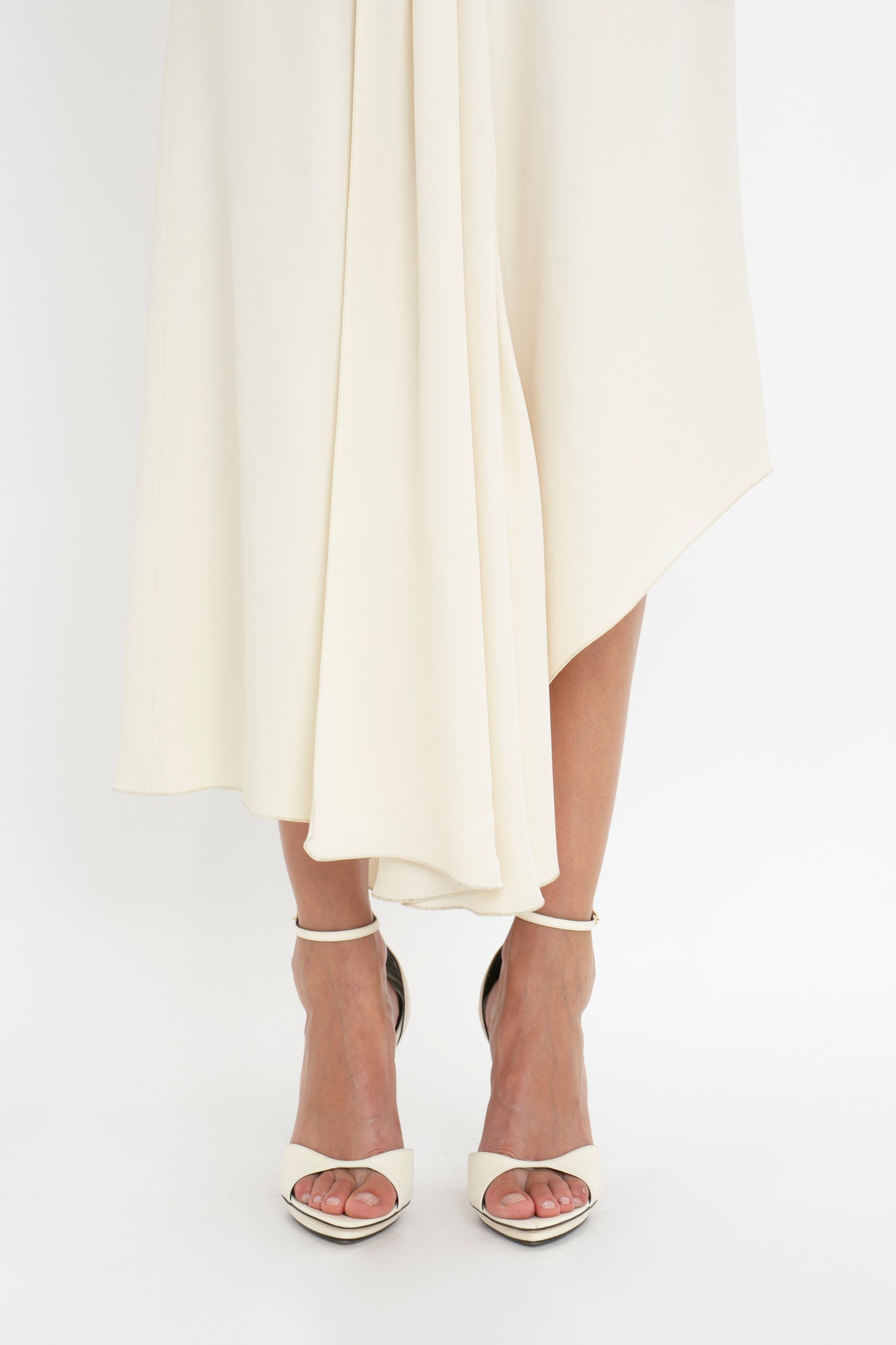 A close-up of a person wearing a flowing, Victoria Beckham Sleeveless Tie Detail Dress In Cream with an asymmetric hemline and white high-heeled sandals, standing against a plain white background.