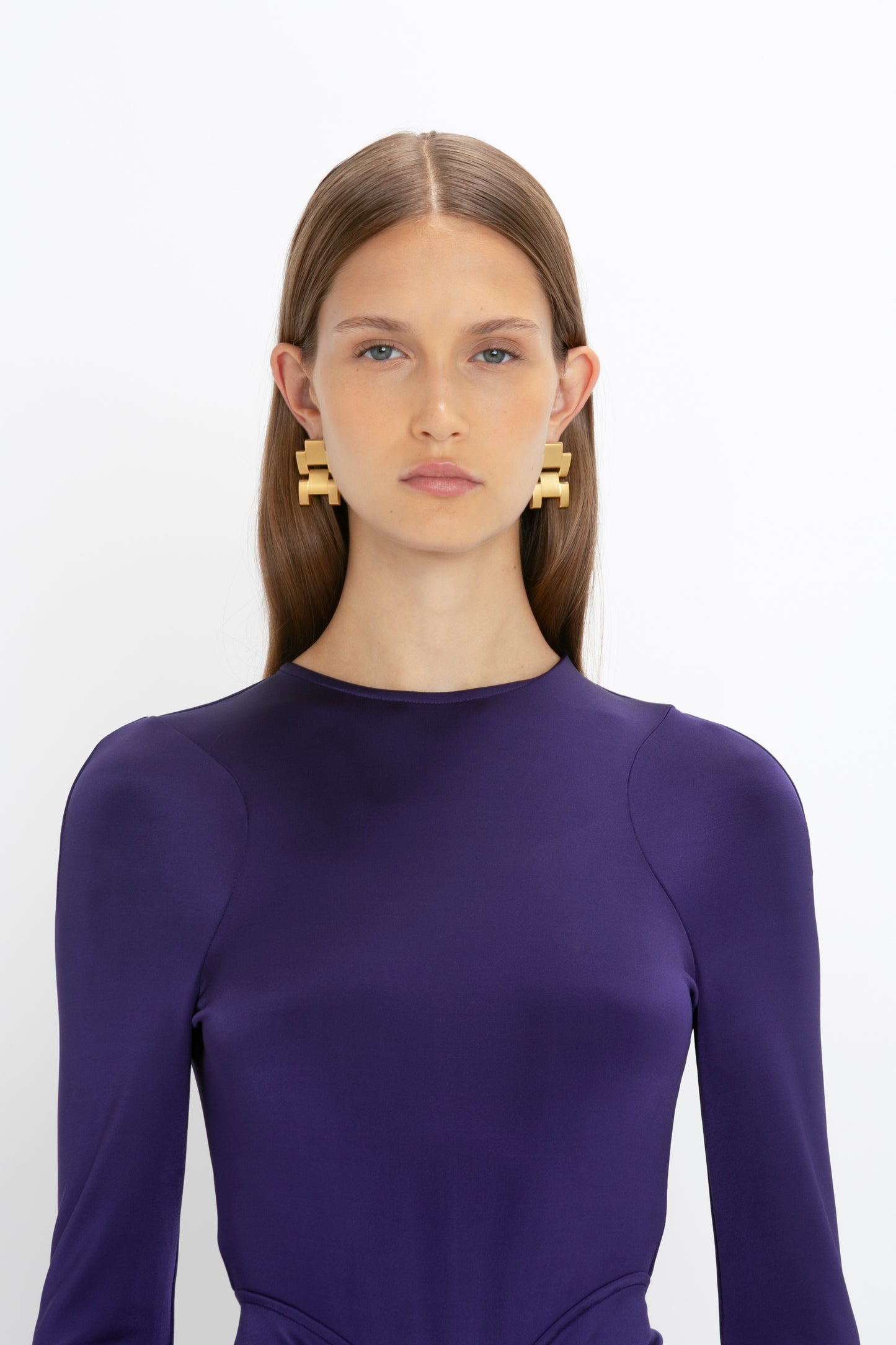 A person with long, straight hair, wearing a Long Sleeve Gathered Midi Dress In Ultraviolet by Victoria Beckham and large, rectangular earrings, stands against a plain white background.