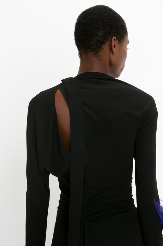 Rear view of a person wearing a Victoria Beckham Slash-Neck Ruched Midi Dress In Black with an asymmetric cut-out, standing against a white background.