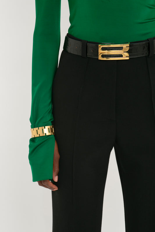 A person wearing black trousers and a green long-sleeve top with a Victoria Beckham Jumbo Frame Belt in Black Croc-Effect Leather and a gold bracelet.