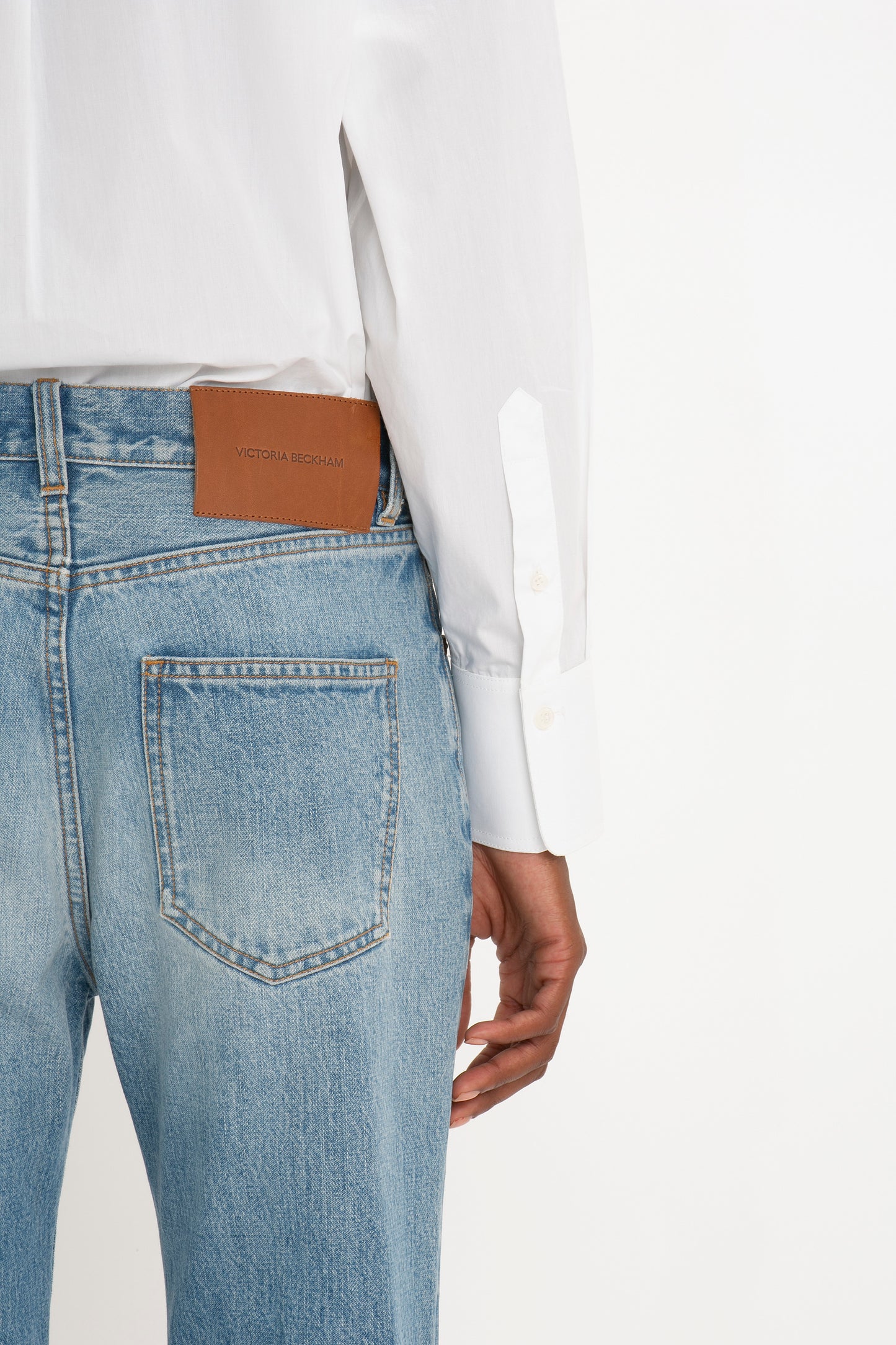 Close-up side view of a person wearing a Victoria Beckham Cropped Long Sleeve Shirt In White tucked into blue jeans, highlighting a brown leather 'Victoria Beckham' branded label on the jeans.