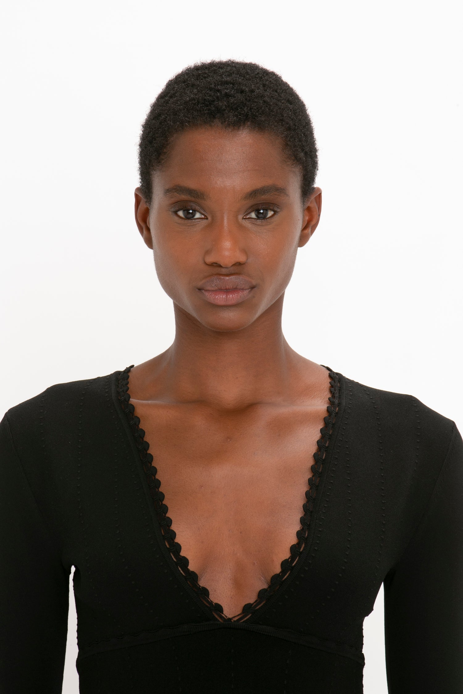 A person with short hair poses in a VB Body Long Sleeve V Neck Dress In Black by Victoria Beckham against a plain white background.