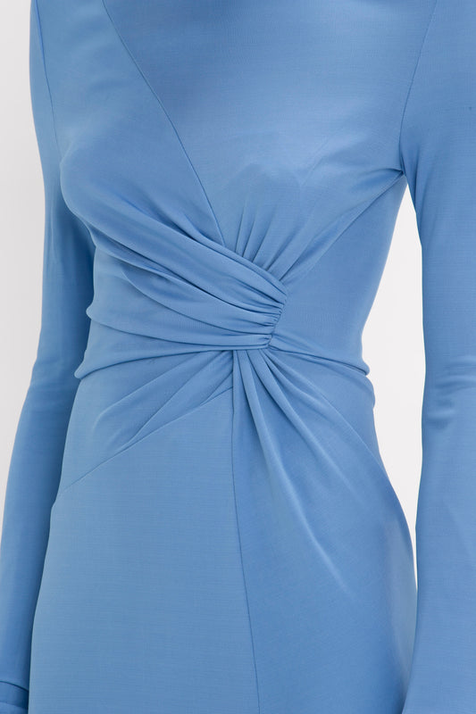 Close-up of a Victoria Beckham High Neck Asymmetric Draped Dress In Oxford Blue with long sleeves and a twisted knot detail at the waist on a gray background.