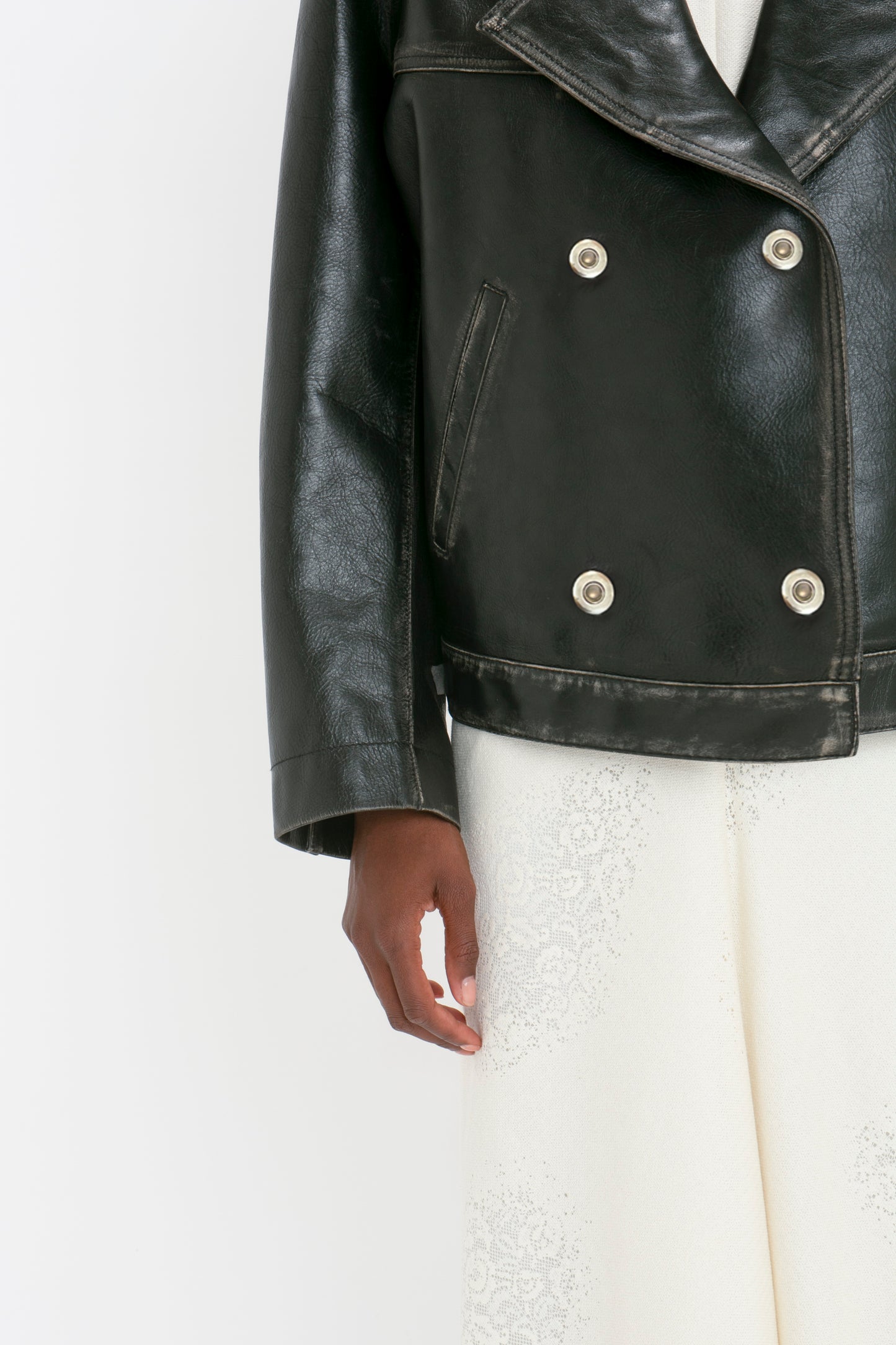 Close-up of a person wearing an Oversized Leather Jacket In Black by Victoria Beckham over a white outfit with lace detailing. Only the bottom half of the jacket, and the person's arm, and a part of the white outfit are visible.