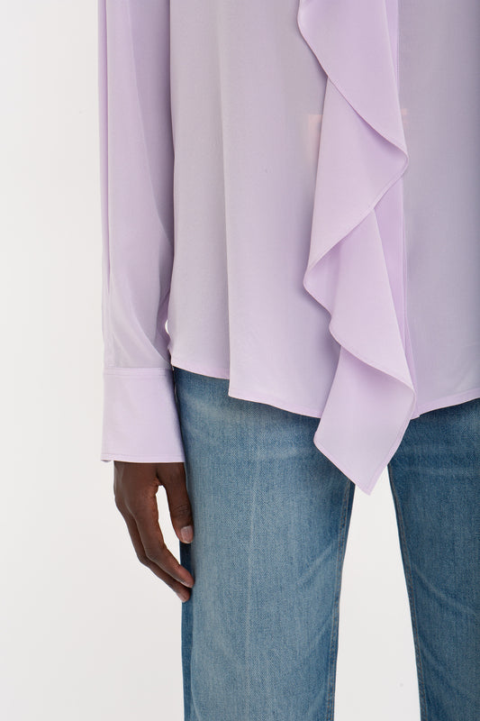 Close-up of a person wearing a Victoria Beckham Asymmetric Ruffle Blouse in Petunia and blue jeans, focusing on the arm and torso against a white background.