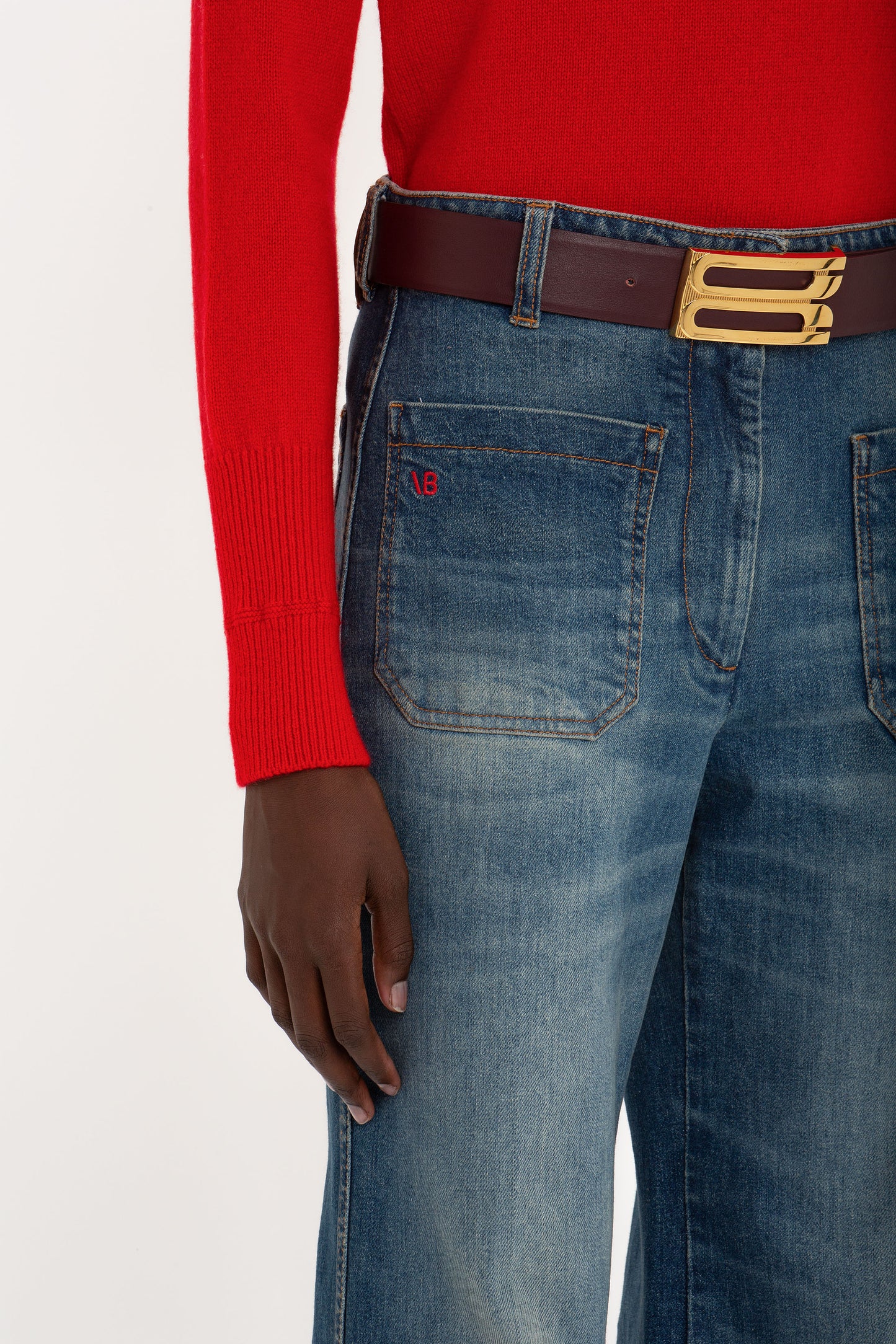 A person wearing a red sweater and blue jeans with a brown belt featuring a large gold buckle. The sweater's sleeve ends mid-hand, and a small red logo appears on the front pocket of the tailored Victoria Beckham Alina High Waisted Jean In Heavy Vintage Indigo Wash.