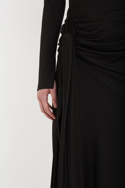Close-up of a woman's side, focusing on her hand gently touching the fabric of her elegant black Victoria Beckham high neck asymmetric draped dress.
