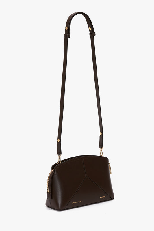 Introducing the Exclusive Victoria Crossbody Bag In Brown Leather, a dark brown calf leather bag with gold accents and a long adjustable strap by Victoria Beckham. The bag features a zippered top closure and a structured, geometric design, perfect for any occasion.