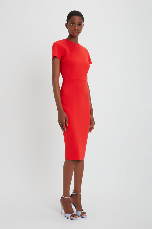A young black woman wearing a Victoria Beckham bright red fitted t-shirt dress with a midi-length hem and black pointy toe stilettos, standing against a white background.