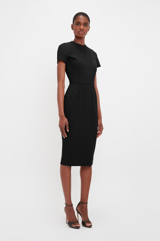 A woman standing in a studio, wearing an elegant Victoria Beckham black knee-length dress with short sleeves and peplum detail, crafted from matte bonded crepe, paired with black heels.