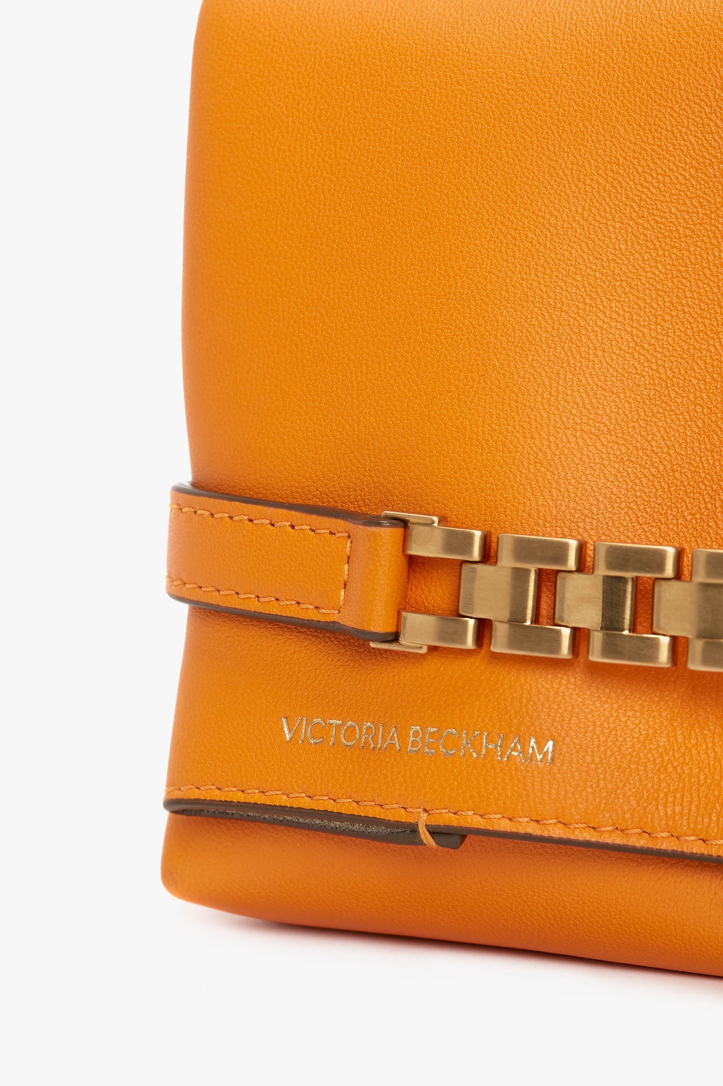 Close-up of an orange **Mini Chain Pouch Bag With Long Strap In Mandarin Leather** featuring gold-tone hardware and a long strap with chain detail. The bag's bright mandarin hue is complemented by the brand name embossed in silver on the leather: **Victoria Beckham UK**.