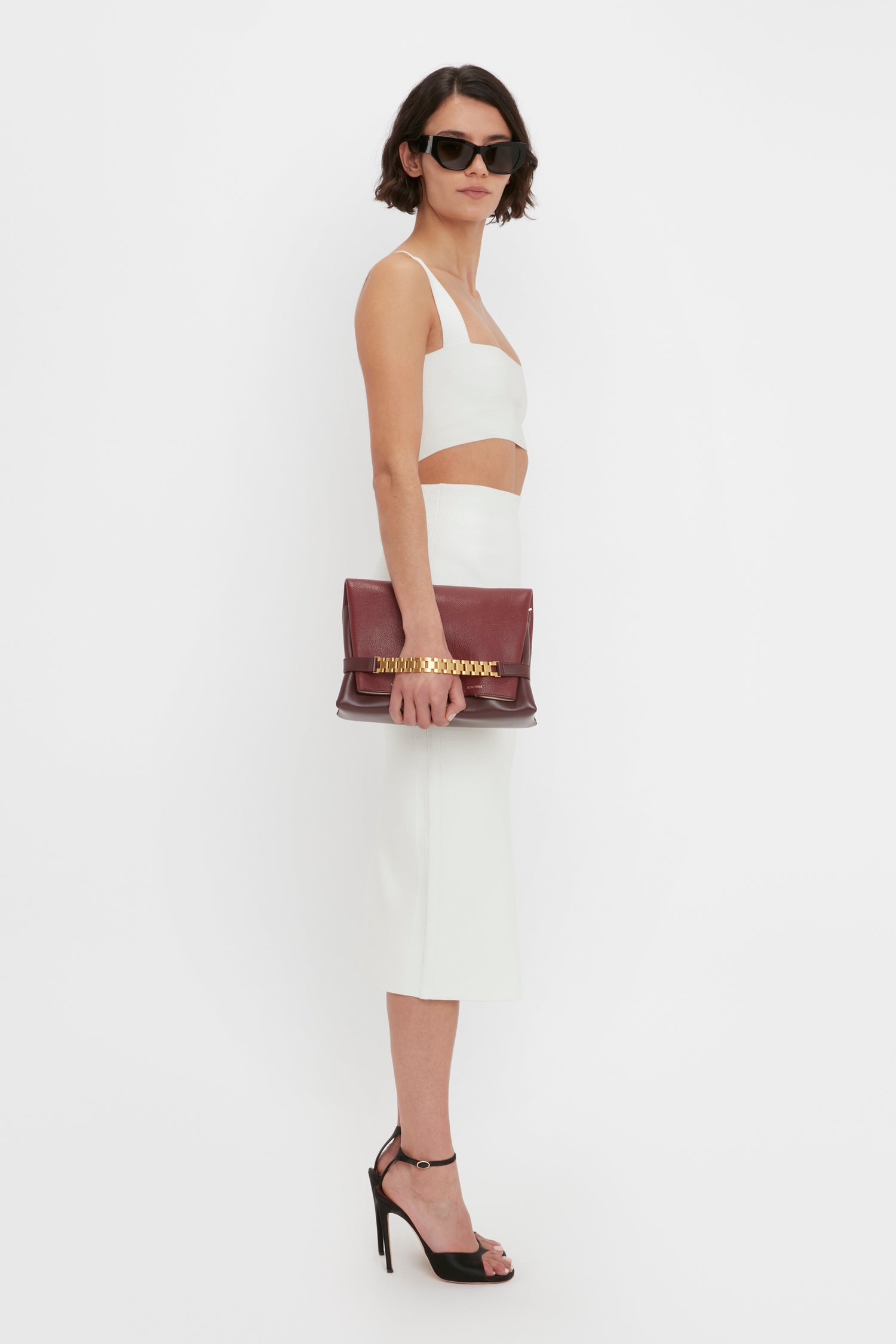 A woman in a chic Victoria Beckham Body Strap Bandeau Top In White and sunglasses, carrying a maroon handbag and wearing black heels, poses against a plain white background.