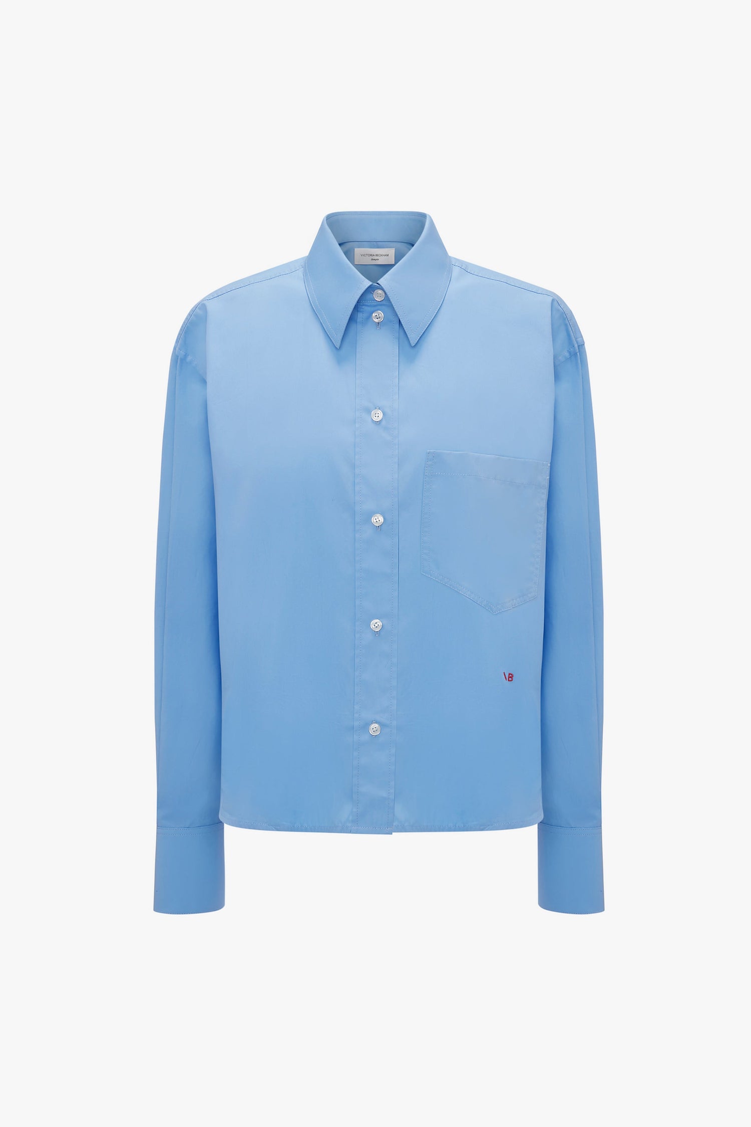 A Cropped Long Sleeve Shirt In Oxford Blue featuring a breast pocket and a small embroidered Victoria Beckham monogram near the pocket's bottom-right corner.
