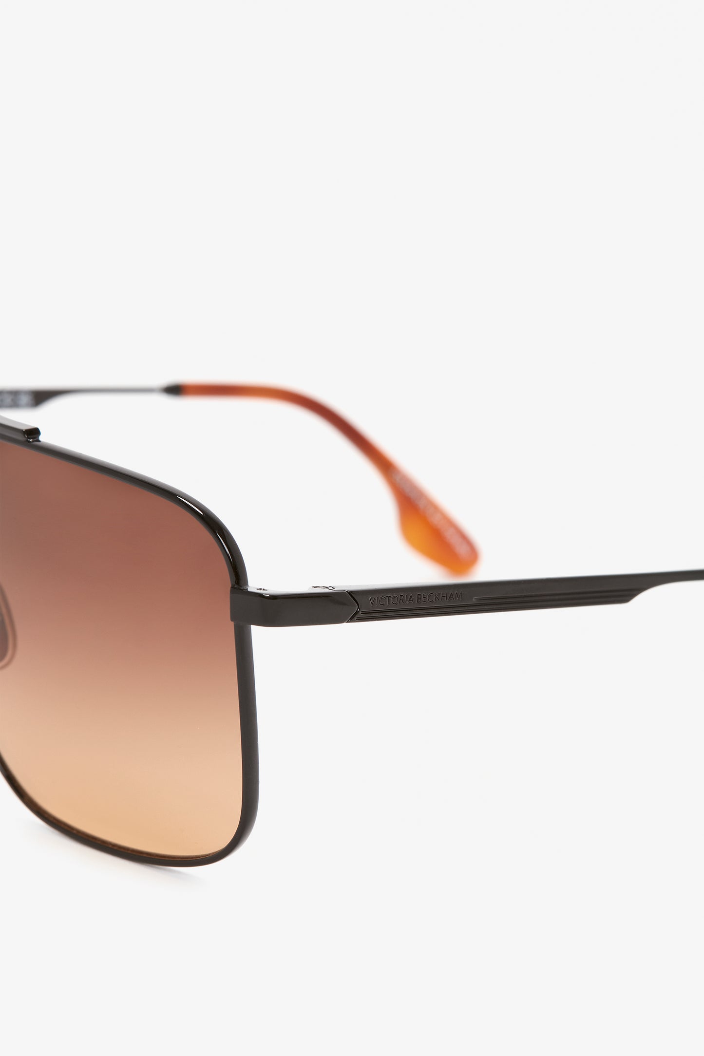 Close-up of a pair of Victoria Beckham Double Bridge V-Line Navigator In Gold-Black-Brown sunglasses with blush gradient lenses, black frame, and orange temple tips against a plain white background.