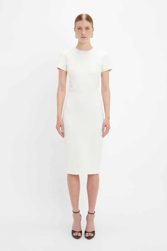 A woman in a Victoria Beckham sleek white fitted T-shirt dress and black sandals stands against a plain white background, looking directly at the camera.