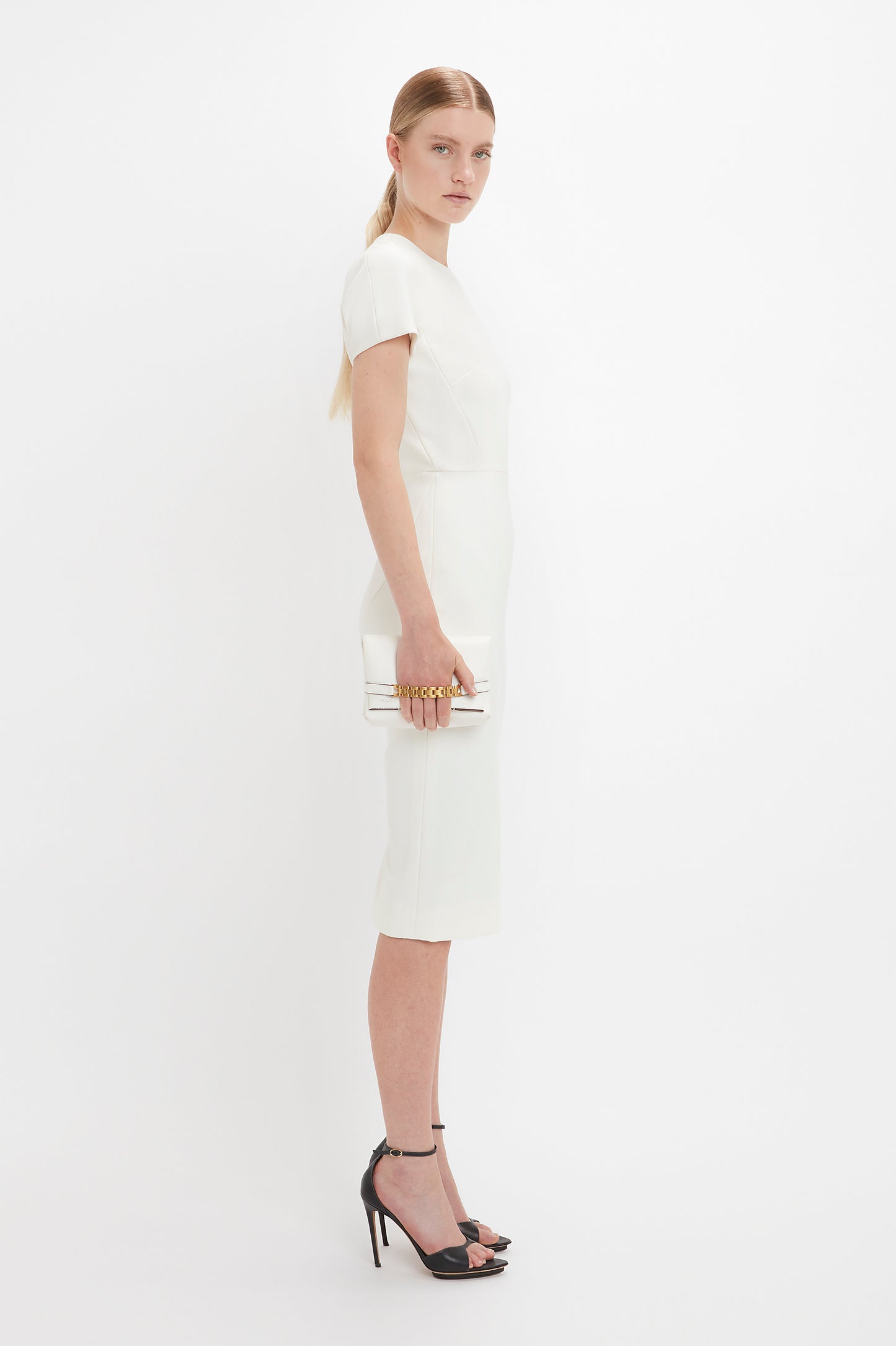 A woman in a sleek white Victoria Beckham fitted T-shirt dress and black pointy-toe stiletto sandals, holding a white mini chain pouch, posing against a plain white background.