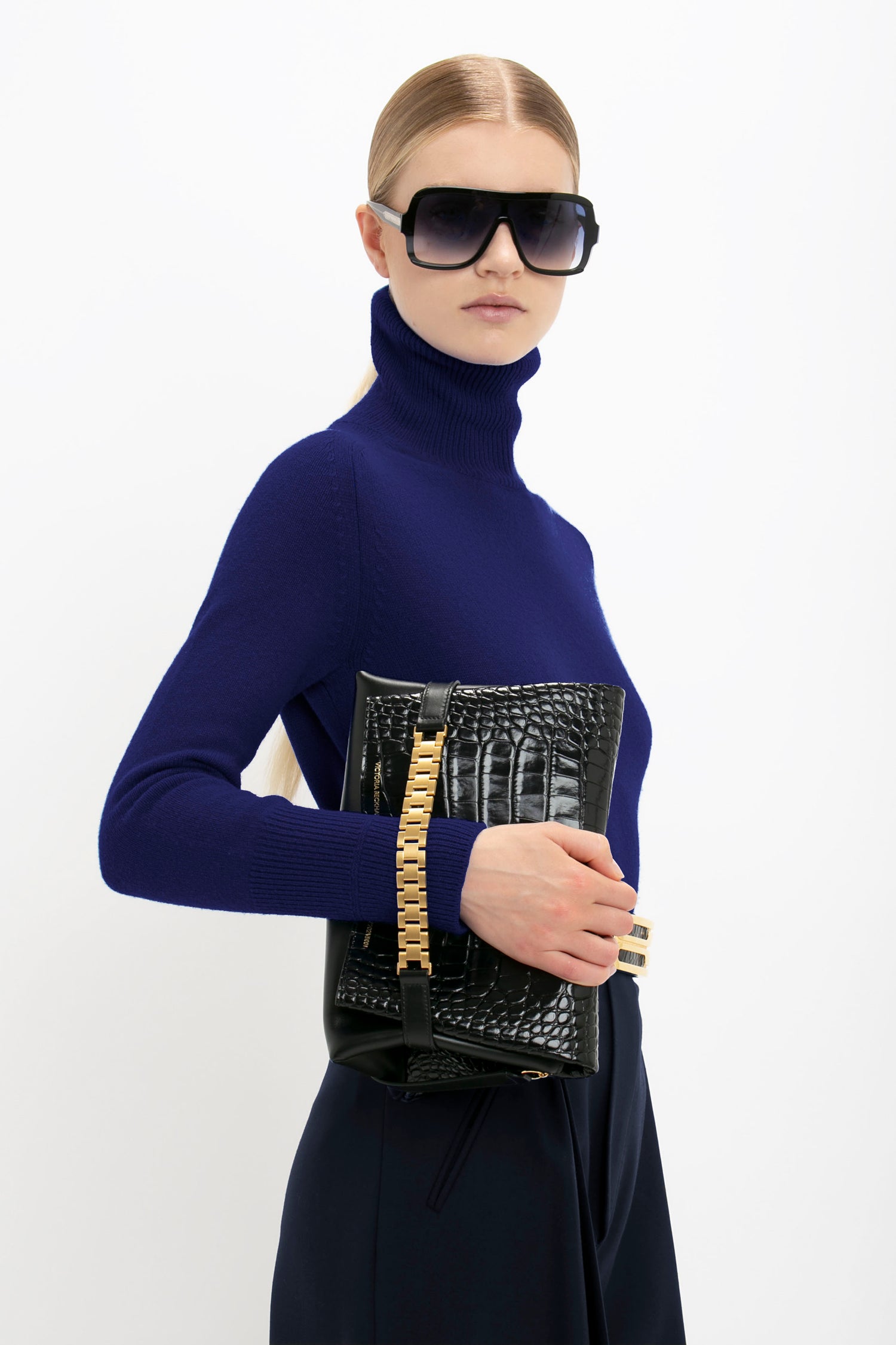 A person wearing a Victoria Beckham Polo Neck Jumper In Navy, dark sunglasses, and holding a black croc-embossed clutch with gold accents evokes a touch of Victoria Beckham's iconic style.