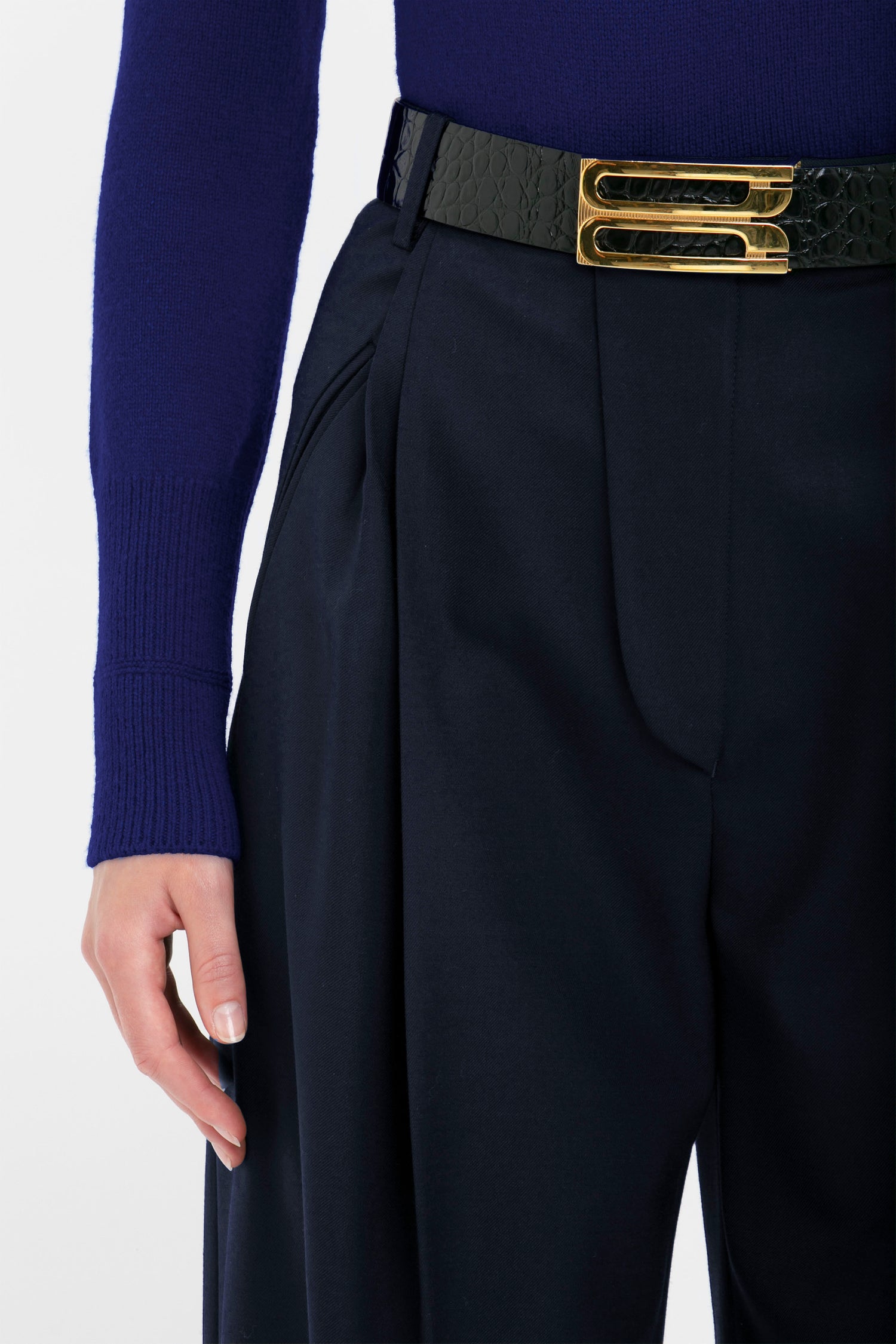 A person wearing a navy blue Polo Neck Jumper In Navy and navy blue pleated trousers with a black belt featuring a double gold buckle. Only the lower part of the textured knit sweater, designed by Victoria Beckham, the trousers, and one hand is visible.