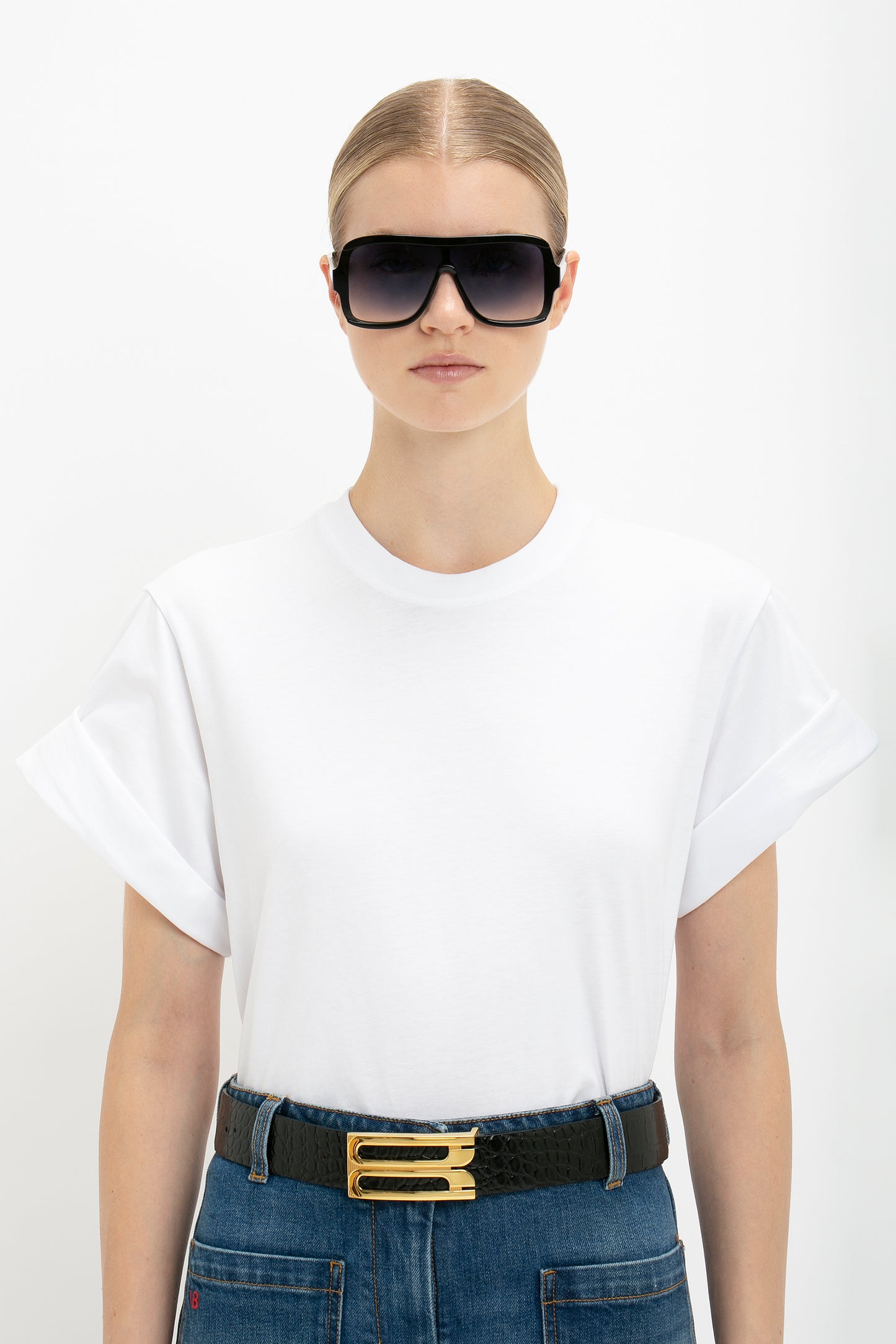 Person wearing an oversized Victoria Beckham Asymmetric Relaxed Fit T-Shirt In White, blue jeans with a belt, and large black sunglasses, standing against a plain white background.
