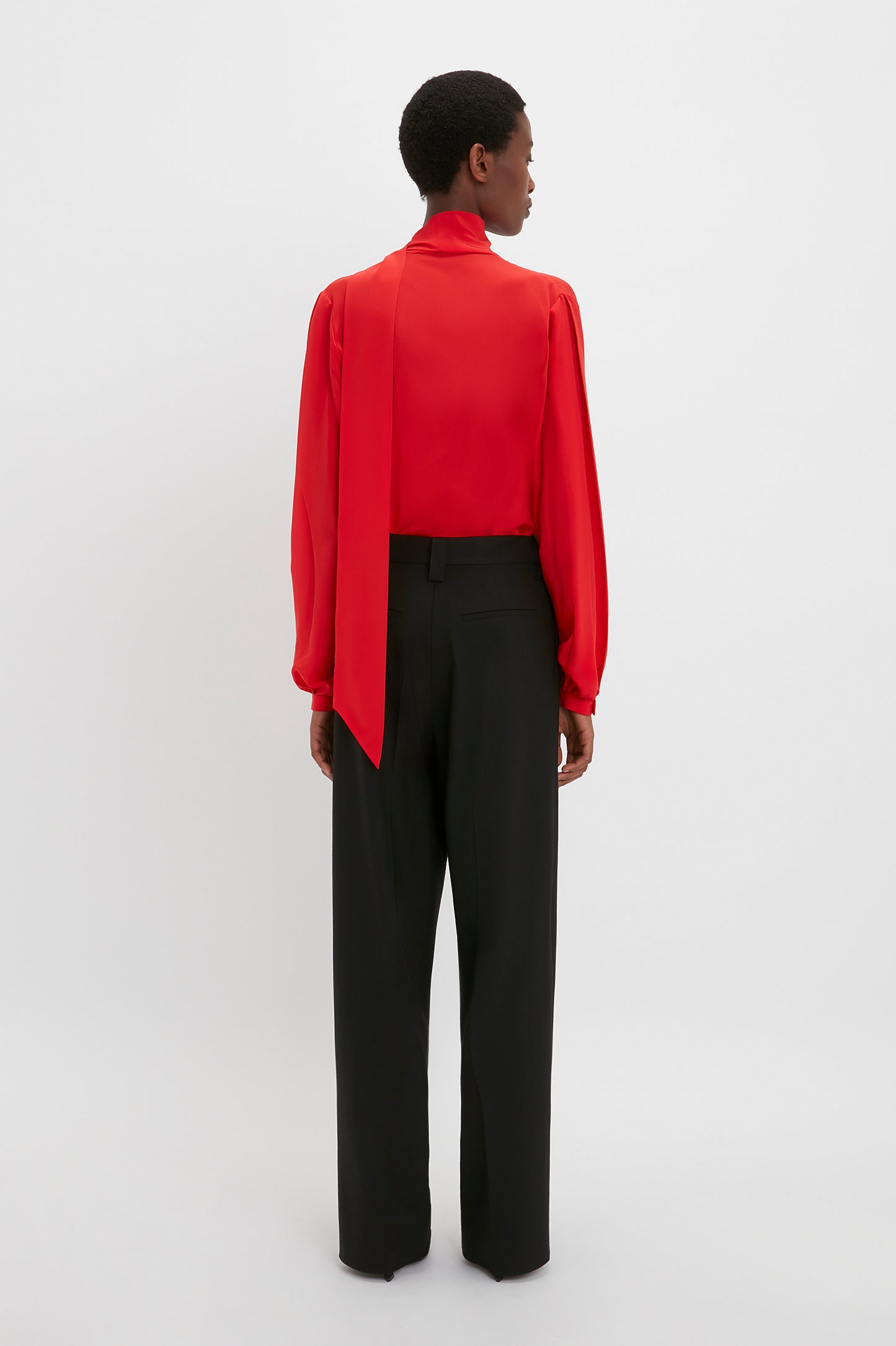 A person is standing with their back to the camera, wearing a Victoria Beckham Scarf Neck Blouse In Bright Red paired with black trousers against a white background.