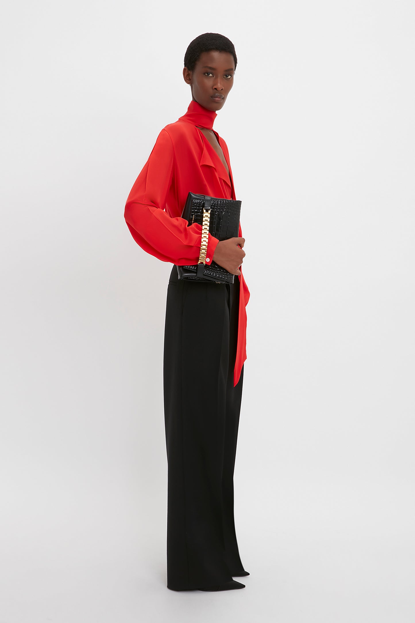 A person stands against a white background wearing a Victoria Beckham Scarf Neck Blouse In Bright Red with a deep-V neckline and black pants, holding a black clutch with a gold chain.