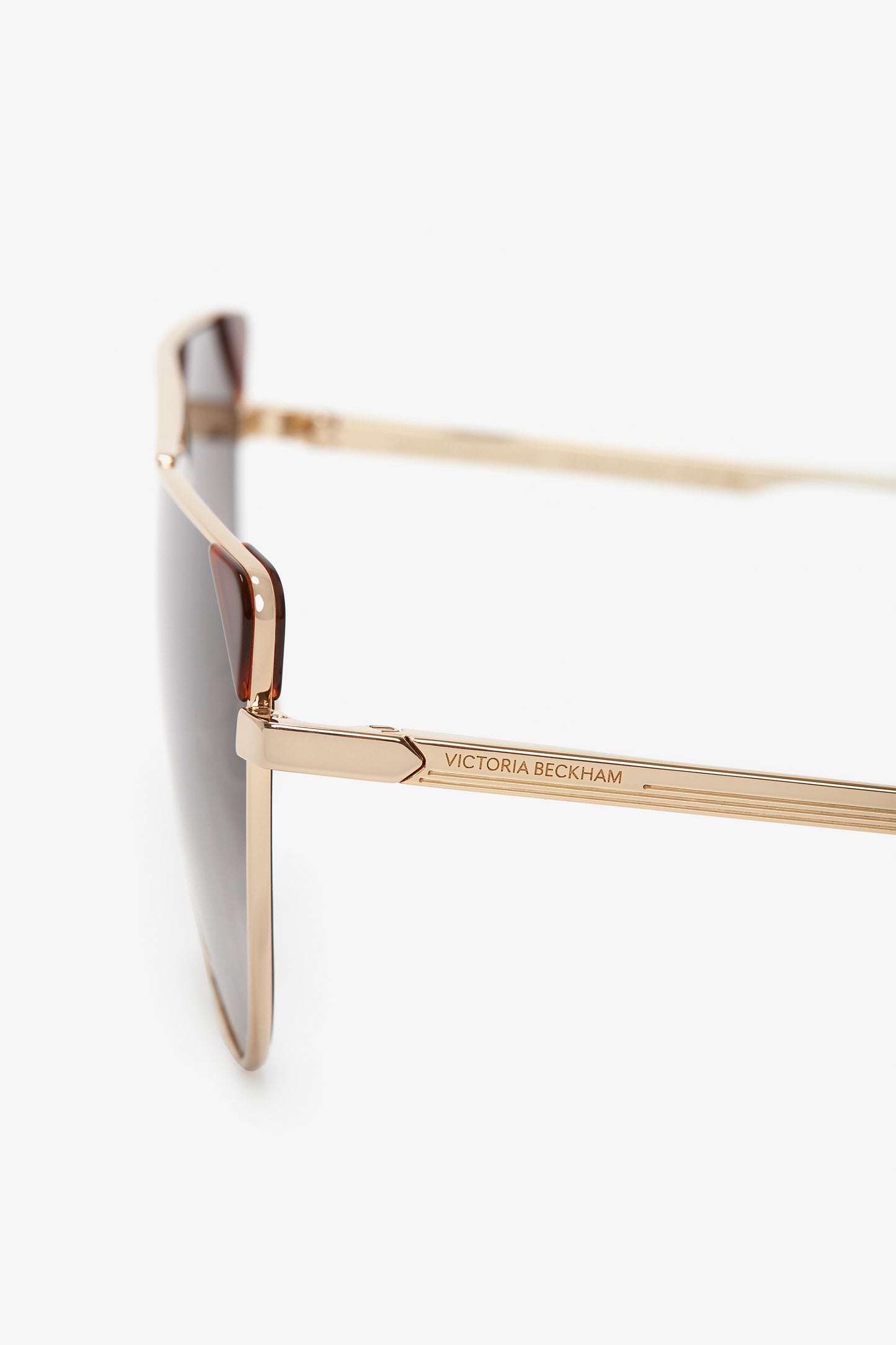 A close-up view of V-Line Aviator In Gold Blonde Havana sunglasses with gold metal frames and dark caramel lenses. The arm is engraved with the name "Victoria Beckham.