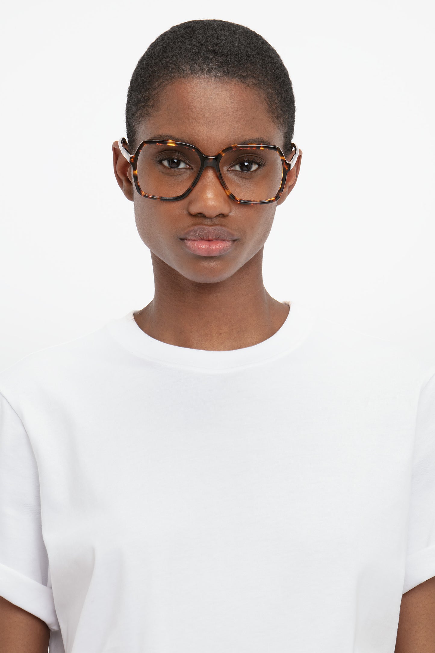 Person with short hair wearing glasses from the Victoria Beckham collection, featuring Guilloche Frame Opticals In Dark Havana tortoise acetate, looking directly at the camera in a plain white t-shirt.