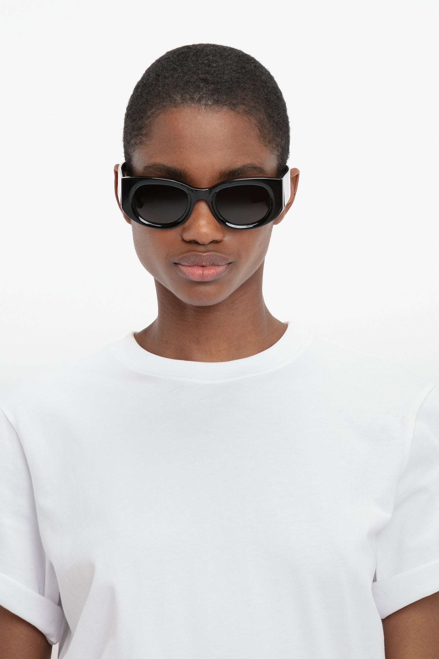 Person with short hair wearing Monogram Detail Sunglasses In Black by Victoria Beckham and a white t-shirt, looking directly at the camera against a white background.