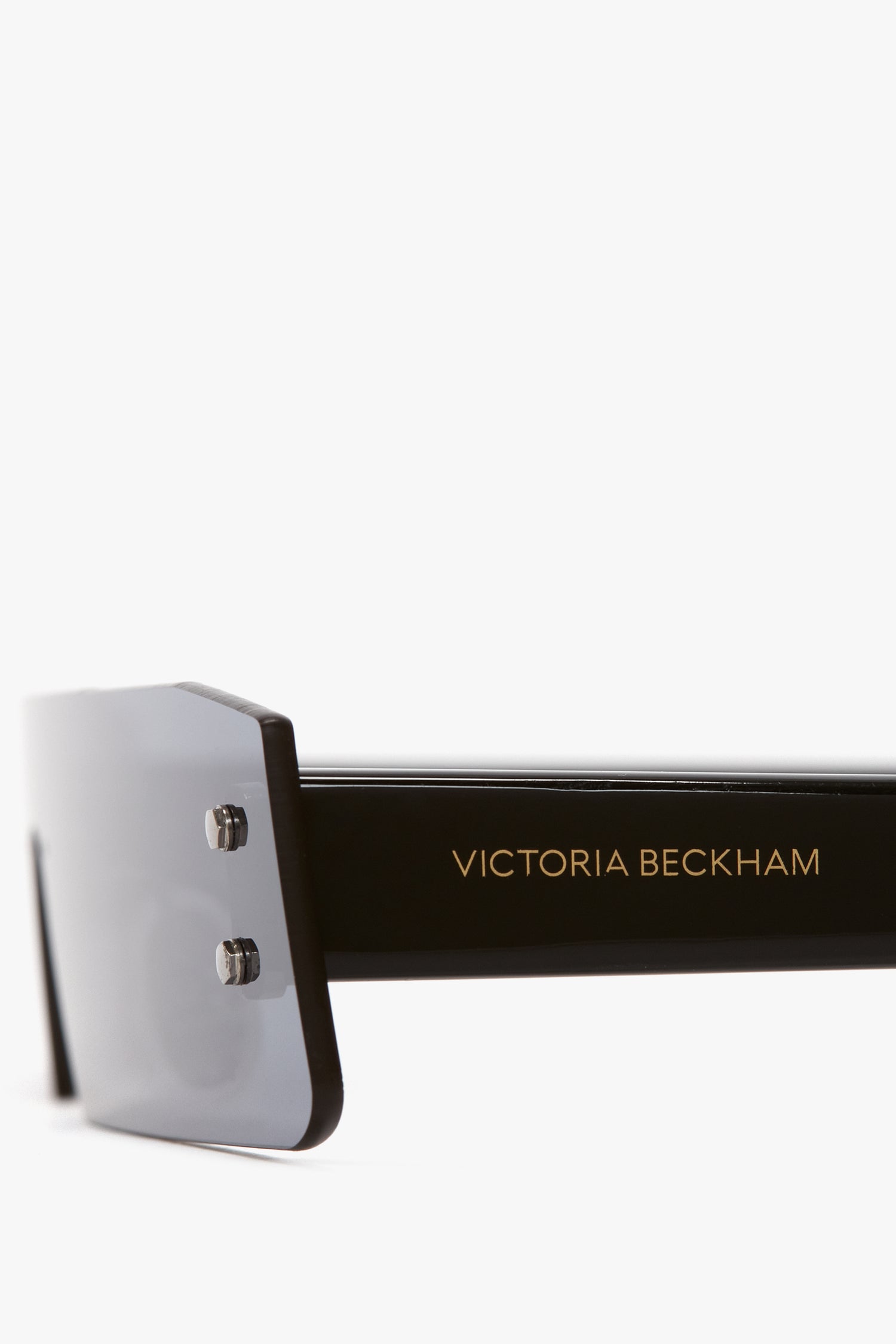 Close-up of black sunglasses with a thick frame and dark blue mirror lenses. The arm of the sunglasses displays the gold text "Victoria Beckham.